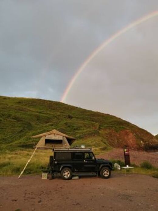 Defender mit Faltdachzelt vor einem Hügel und im Hintergrund ein Regenbogen