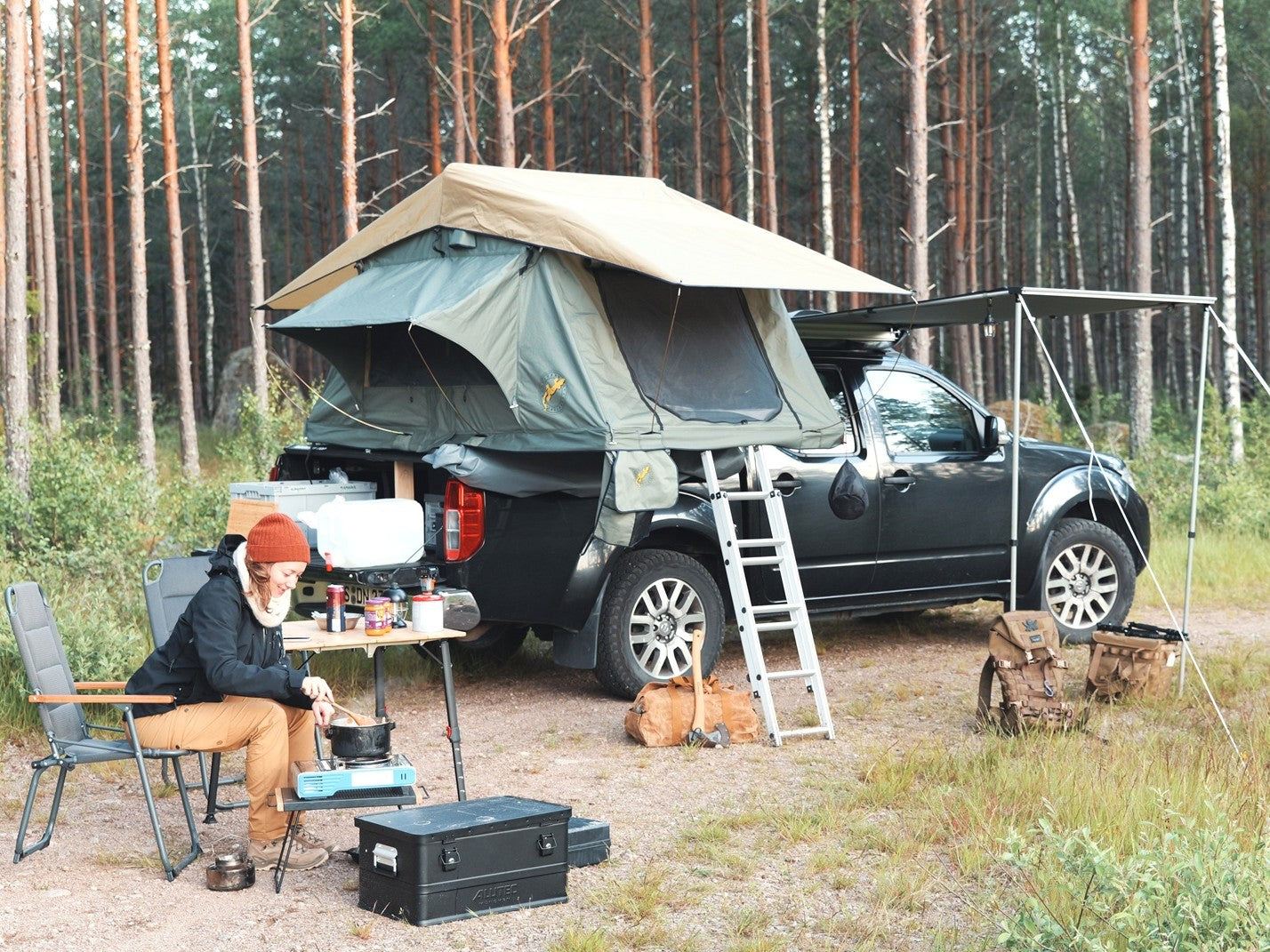 Pickup mit Dachzelt und Markise - Frau sitzt beim Kochen auf Campingstuhl davor